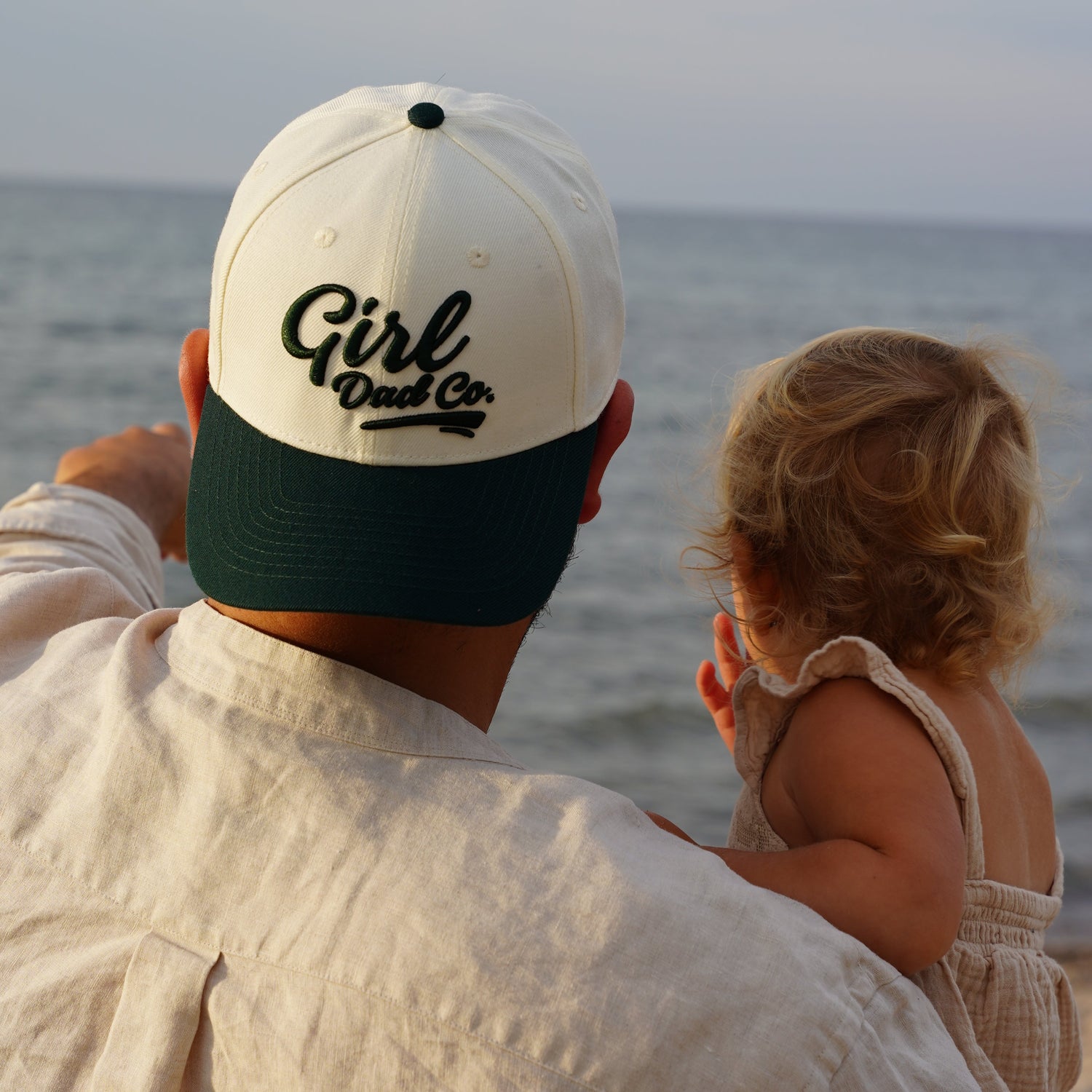 A person wearing a white and green baseball cap with 'Girl Dads' text on it, is seen from the back, looking out at the sea with a young child.