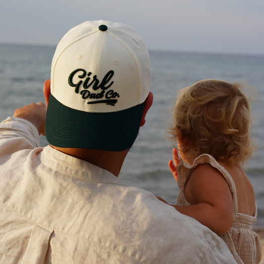 A person wearing a white and green baseball cap with 'Girl Dads' text on it, is seen from the back, looking out at the sea with a young child.